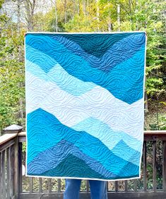 a person holding up a blue and white quilt in front of a wooden deck with trees