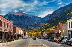 a street lined with parked cars in front of tall mountain range covered in fall foliage