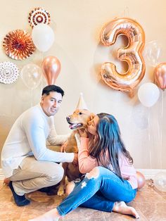 a man and woman sitting on the floor with their dog in front of balloons that read 3