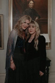 two women standing next to each other in front of a portrait on the wall behind them