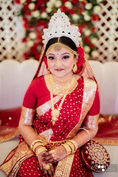 a woman in a red and gold bridal outfit sitting on a white couch with flowers behind her