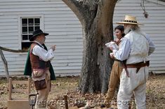 three people dressed in period clothing standing near a tree and bench, talking to each other