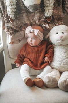 a baby sitting on a chair next to two stuffed animals and one is wearing a sweater