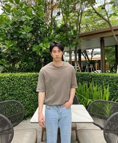 a young man standing in front of a table with chairs around it and bushes behind him