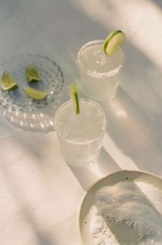 two glasses filled with water and limes on top of a white table covered in powder