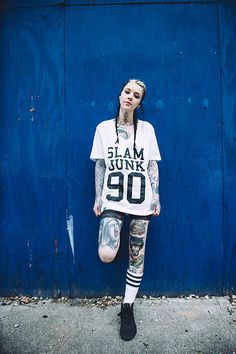 a woman with tattoos standing in front of a blue wall wearing a t - shirt that says slam junk 90