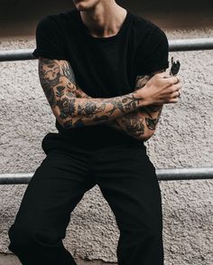 a man with tattoos is leaning against a fence and holding his arms crossed, looking at the camera