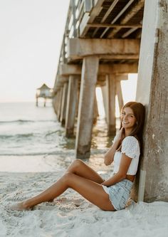 a beautiful young woman sitting on top of a sandy beach next to a wooden pier