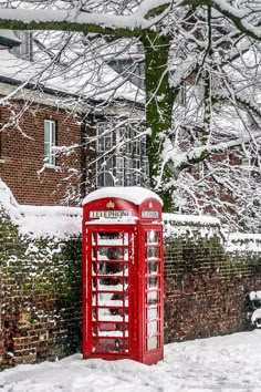 a red phone booth sitting in the snow next to a brick wall and tree with no leaves on it