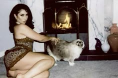 a woman kneeling down next to a cat in front of a fire place