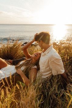 a man and woman laying in tall grass near the ocean with their eyes closed as the sun goes down