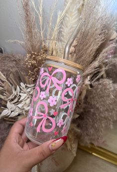 a hand holding a cup with pink flowers and bows painted on the side, in front of some dried grass