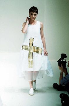 a woman is walking down the runway wearing a white dress with a cross on it