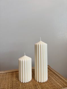 two white candles sitting next to each other on a bamboo mat in front of a gray wall