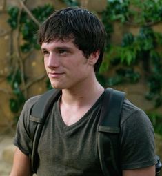 a young man with a backpack standing in front of a stone wall and looking at the camera