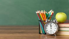 an alarm clock sitting on top of a stack of books next to an apple and pencils