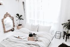 a bedroom with white walls and flooring has a large bed, mirror, plants, and books on it