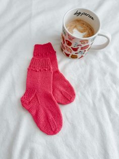 a cup of coffee next to a pair of pink knitted mitts on a bed