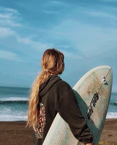 a woman holding a surfboard on top of a beach next to the ocean and blue sky