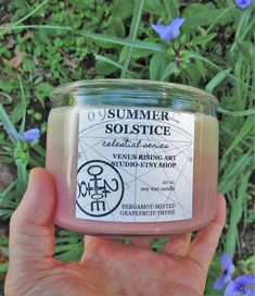 someone holding up a jar of soap in front of some blue and green flowers with the label summer solstice on it