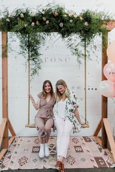 two women sitting on a swing in front of a wall with flowers and greenery