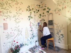 a woman sitting on a chair painting a wall with flowers and plants painted on it