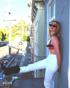 a woman leaning against the side of a building while wearing white pants and black boots