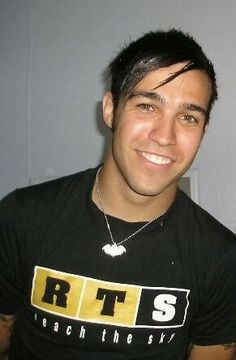 a man wearing a black tshirt smiles at the camera while standing in front of a wall