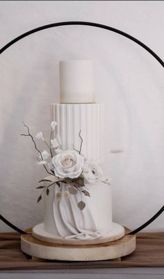 a wedding cake with white flowers and greenery sits on a wooden stand in front of a circular frame