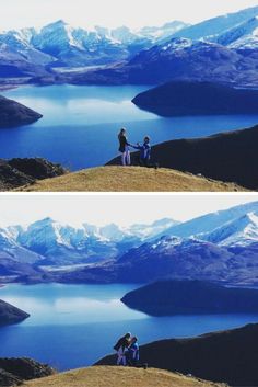 two people sitting on top of a hill next to a body of water with snow covered mountains in the background