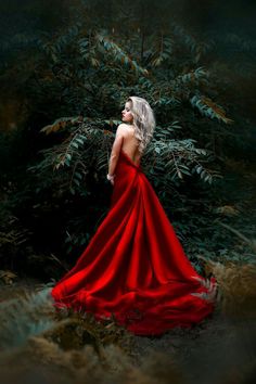 a woman in a red dress is standing by some bushes and trees with her back turned to the camera