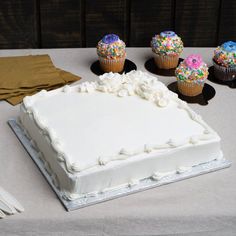 a white frosted cake sitting on top of a table next to cupcakes