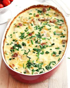 a casserole dish with meat and vegetables in it on a wooden table next to a bowl of tomatoes