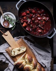 a wooden cutting board topped with bread next to a pot of stew