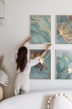 a woman is hanging paintings on the wall in her living room with white furniture and gold accents