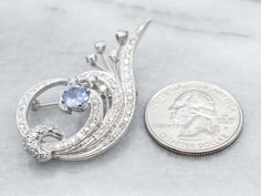 This vintage sapphire and diamond brooch is fit for a queen, crafted of luxurious materials, and completed with immaculate workmanship. This is a retro-era piece crafted of brilliant, polished 14-karat white with glittering diamonds set in a sweeping botanical motif. We've finished this exquisite vintage brooch with a sapphire worthy of the setting. This stone is the perfect shade of periwinkle blue, lustrous and crystal clear. Love this piece, but want to make it more versatile? We here at Market Square Jewelers and our master metalsmiths can convert this fantastic brooch into a necklace or pendant for an additional fee. Please contact us for more information! Metal: 14K White GoldGem: Sapphire 1.10 CaratsGem Measurements: 6.1 mm, RoundAccents: Diamonds totaling 1.39 Carats, VS-I in Clari Elegant Silver Sapphire Brooches, Diamond Brooch With Brilliant Cut, Fine Jewelry Diamond Brooch With Brilliant Cut, Diamond Brooches With Brilliant Cut In Fine Jewelry Style, Elegant Sapphire Brooches For Formal Occasions, Elegant Sapphire Brooch Jewelry, Elegant Diamond Gemstone Brooches, Retro Era, Market Square