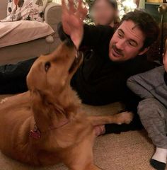 a man sitting on the floor next to a little boy and a brown dog with its mouth open