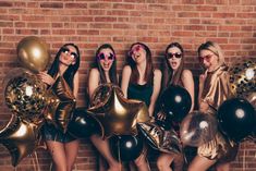 four girls in gold dresses and sunglasses are posing with black and gold balloons against a brick wall