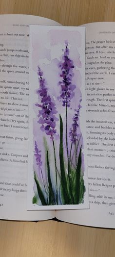 an open book with watercolors on it and purple flowers in the middle, sitting on a wooden table