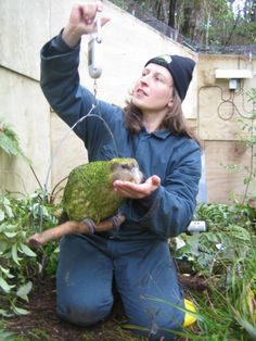 a woman holding a bird in her hand while sitting on the ground next to plants