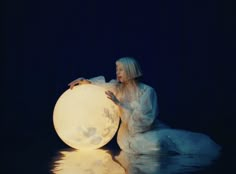 a woman is sitting on the ground next to a large light ball in the water