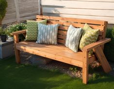 a wooden bench with four pillows on it in the grass next to a fence and shrubbery