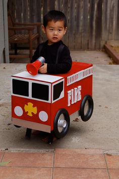 a little boy sitting on top of a firetruck shaped cardboard toy car in front of a wooden fence