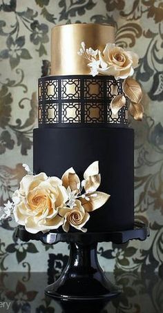 a black and gold wedding cake with white flowers on the bottom tier is displayed against a floral wallpaper