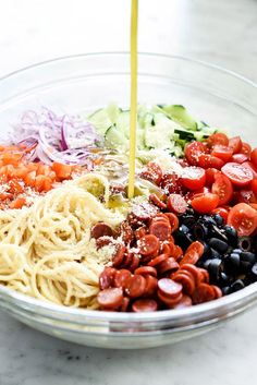 a bowl filled with pasta, olives, tomatoes, and other ingredients being poured into it