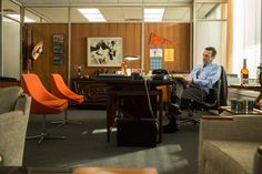 a man sitting at a desk in an office with orange chairs and a painting on the wall