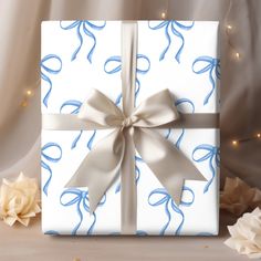 a white gift box with a blue ribbon and bow on it sitting in front of some flowers