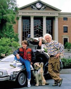 two men and a dog sitting on the back of a car in front of a building