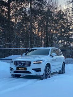 a white volvo suv is parked in the snow near some trees and fenced area