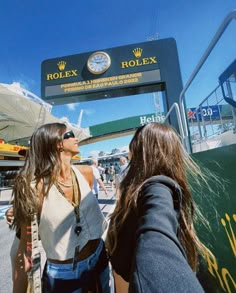 two women standing in front of a rolex sign at an outdoor event on a sunny day